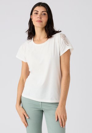 Mujer con cabello oscuro hasta los hombros, vestida con una camisa blanca de manga corta con detalle de encaje en los hombros y pantalones verde claro, de pie frente a un fondo gris.