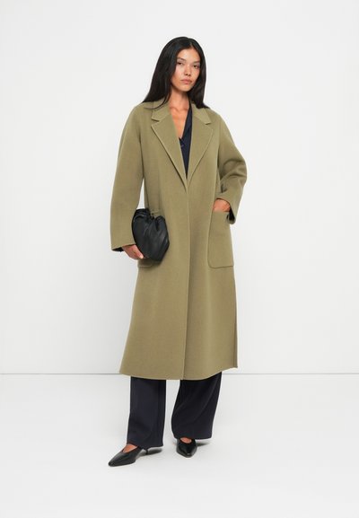 Cappotto di lana verde oliva con colletto a revers, taglio oversize, tasche laterali e maniche lunghe, abbinato a pantaloni scuri e una borsa nera.