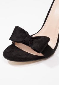 Talons ouverts en daim noir avec un détail de nœud texturé sur la bride avant et une couture beige visible sur la semelle intérieure.