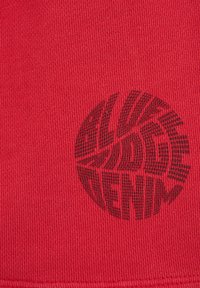 Červená tkanina s texturovaným povrchem, na které se nachází velké černé kruhové logo s stylizovaným textem "ALWAYS DENIM" v tučném písmu.