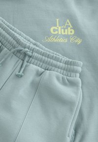 Vaaleansininen urheiluasusetti, johon kuuluu collegepaita ja juoksuhousut, joissa on joustava vyötärönauha ja kiristysnyöri. "LA Club Athletics City" keltaisella painatuksella.