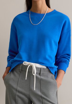 Personne portant une chemise bleu vif à manches longues, un pantalon gris avec un cordon blanc et un collier en chaîne argentée, mains dans les poches.