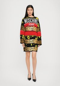 Moteris vilkinti ilgomis rankovėmis megztinį su suknele, ant kurios yra užrašas "MOSCHINO JEANS", raudonos, juodos, geltonos spalvų ir tigro rašto motyvai, taip pat juodos smailianos įkapių.