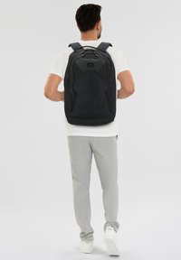 Zaino nero con un design strutturato, numerose tasche e spallacci imbottiti. Realizzato in tessuto resistente, con un logo sulla parte frontale.