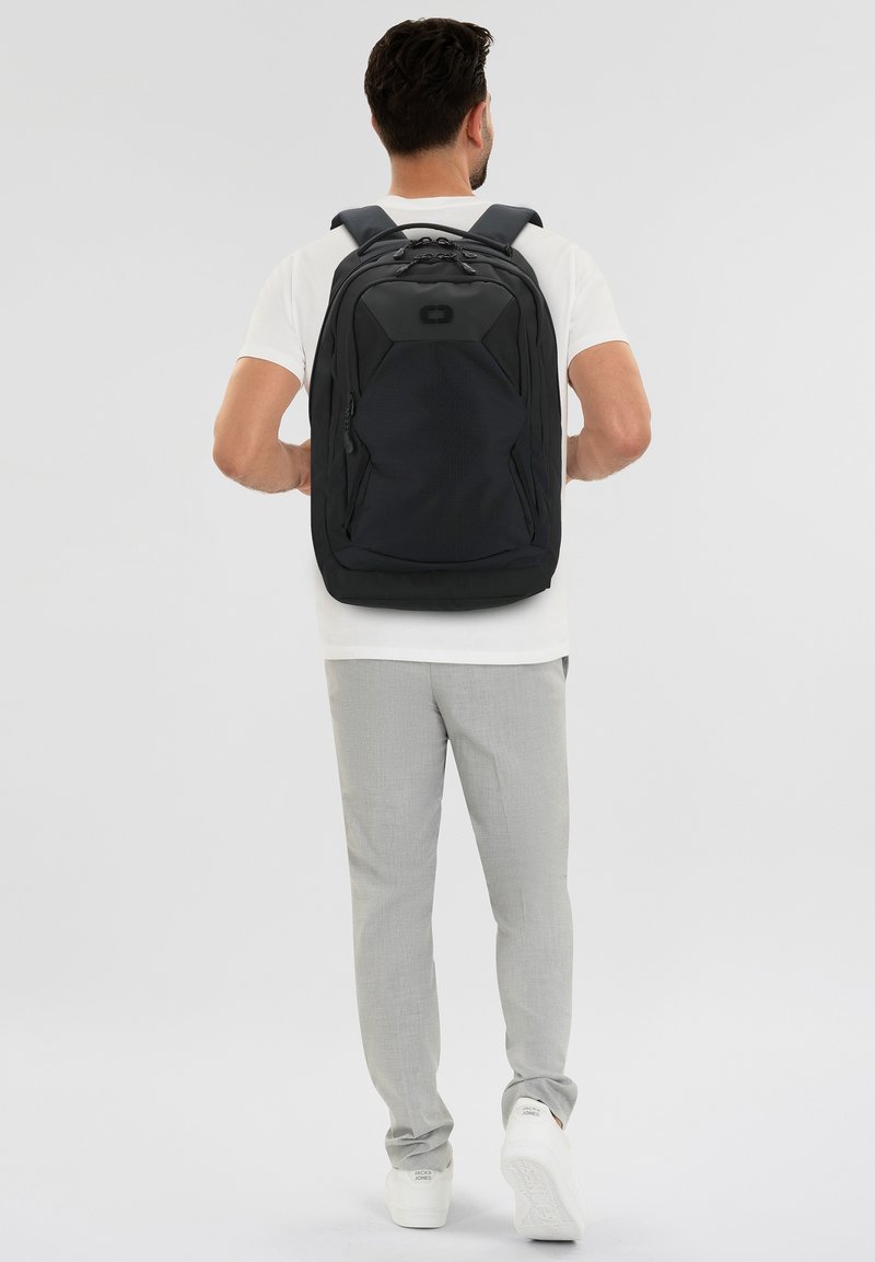 Zaino nero con un design strutturato, numerose tasche e spallacci imbottiti. Realizzato in tessuto resistente, con un logo sulla parte frontale.