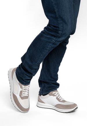 Beine, die dunkelblaue Jeans und weiße Turnschuhe mit beigen Wildlederdetails tragen, stehen mit einem Fuß über dem anderen auf weißem Hintergrund.