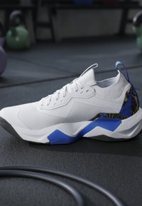 Sneaker atletica bianca con accenti blu e neri posata su un pavimento da palestra con kettlebell e corda per esercizi sullo sfondo.