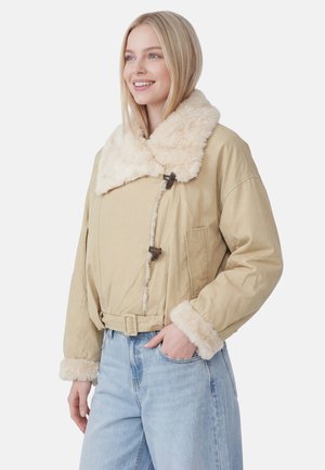 Ung kvinde smiler, iført en beige lammeskindsjakke med stor krave og manchetter, kombineret med lyseblå jeans, stående foran hvid baggrund.