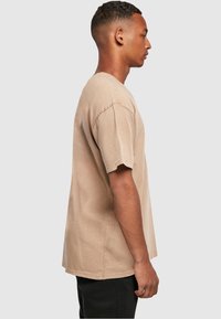 Young man standing in profile wearing a beige oversized short-sleeve t-shirt and black pants against plain background.