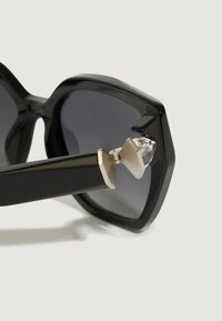 Solbriller med overdimensjonerte svarte heksagonale rammer, grå tonede linser og metallisk hengsel-detall med en krystallakssent.