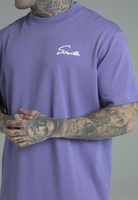 Lilla bomulds t-shirt med rund hals og korte ærmer, prydet med et hvidt kursiv logo på venstre bryst. Textureret stof og afslappet pasform.