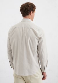 Hellbeige Hemd mit dünnen weißen Streifen, knöpfbarem Kragen und langen Ärmeln. Der Rücken hat einen Schulterbesatz und einen leicht abgerundeten Saum.