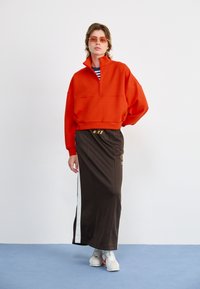 Rød zip-up hettegenser, brun skirt med hvit side stripe og Nike-logo, hvite joggesko, person med oransje solbriller.