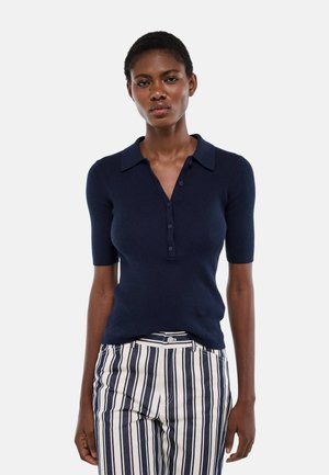 Femme portant un polo bleu marine à manches courtes et boutons, ainsi qu'un pantalon blanc taille haute avec des rayures verticales bleu marine, debout devant un fond blanc.