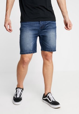 Shorts en denim d'un bleu moyen, coupe droite, avec un ourlet effiloché. Associés à des baskets noires avec des lacets blancs et des accents latéraux.