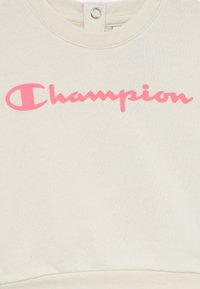 Tissu crème avec un logo "Champion" rose en écriture cursive sur la poitrine et une fermeture à pression au niveau du col.