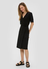 Robe noire avec un décolleté en V profond, manches courtes et ceinture à la taille. Tissu léger avec une texture lisse.