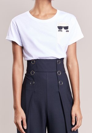 Chemise blanche à manches courtes avec un dessin de visage cartoon portant des lunettes de soleil sur la poche, associée à un pantalon taille haute bleu marine avec des boutons anneaux dorés.