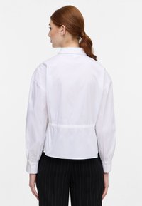 Camicia bianca con bottoni e vita strizzata, maniche lunghe e colletto classico, abbinata a pantaloni neri a righe verticali. Texture liscia.