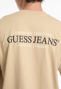 Bežs kokvilnas T-krekls ar tekstu "AMERIKĀNU TRADĪCIJA" virs "GUESS JEANS" un "ORIģINĀLIE DIZAINI" melnā krāsā, kas redzams uz muguras.