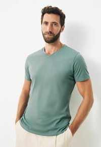 Tricou cu mâneci scurte, de tip v-neck, dintr-un material moale în nuanță de teal. Are un design ajustat și o textură netedă, asortat cu pantaloni de o culoare deschisă.