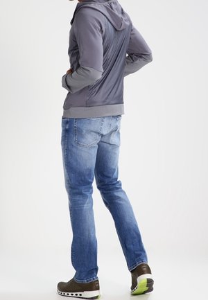 Sudadera gris con cremallera y tejido texturizado, jeans azules y zapatillas oscuras con acentos verdes. Costuras visibles y diseño ajustado.