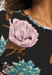 Femme portant un tissu noir avec un grand imprimé floral rose et bleu près de l'encolure et des feuilles brunes.
