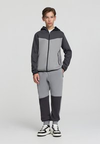 Felpa grigia e nera con zip e cappuccio, caratterizzata da un design color-block. Abbinata a pantaloni coordinati, entrambi realizzati in un tessuto morbido e testurizzato.