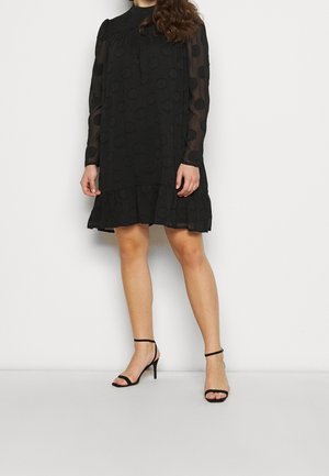 Donna che indossa un vestito nero a maniche lunghe con maniche trasparenti a pois e orlo arricciato, abbinato a tacchi neri open toe con cinturino alla caviglia.