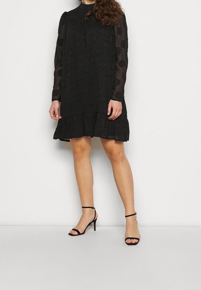 Femme portant une robe noire à manches longues avec des manches transparentes à pois et un ourlet volanté, associée à des talons noirs ouverts avec bride à la cheville.