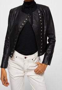 Musta nahkatakki, jossa pyöreä kaulus, koristeelliset hopeiset niitit edessä ja istuvat hihat. Päällä musta paita.