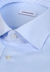 Camicia azzurro chiaro con tessuto strutturato, collo a punta e chiusura con un solo bottone. L'etichetta recita "seidensticker."