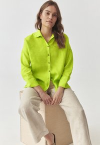 Limonengr�ünes Leinenhemd mit Knöpfen und Kragen, getragen mit cremefarbener Leinenhose. Das Model sitzt auf einer Holzkiste, die Hände im Schoß.