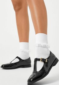 Pantofi T-bar din piele lăcuită neagră, cu finisaj lucios, detalii cu cataramă și cusături decorative, purtați cu șosete albe cu volane.