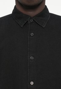 Juodas džinsinis marškinis su klasikine apykakle, šešiais priekinių sagų ir tekstūruotu audiniu. Matomos subtilios siūlės ir sagų akcentai.