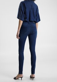 Jeans ajustados de mezclilla azul con cintura alta, con bolsillos traseros y detalles de costura sutiles, combinados con una blusa oscura de corte holgado.