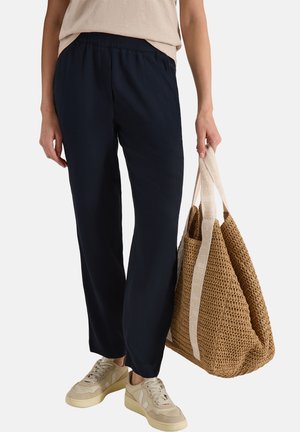 Personne portant un pantalon noir et des baskets beige, tenant un grand sac fourre-tout tissé couleur fauve avec des bretelles claires.