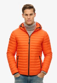 Orange pufferjakke med hætte, der har et quiltet design, to lynlåslommer i siden og en grå ribstrikket turtleneck under.