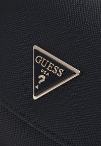 Černý texturovaný povrch se zlatým trojúhelníkovým logem Guess USA, v jehož středu je otazník.