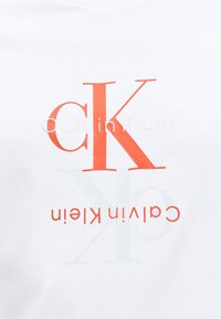 Λευκό ύφασμα με μεγάλα πορτοκαλί γράμματα «cK» και μικρότερο λευκό και πορτοκαλί κείμενο «Calvin Klein», μερικά κείμενα είναι ανάποδα.