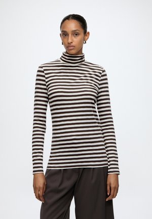Donna che indossa un top a collo alto a righe orizzontali marroni e bianche e pantaloni marroni scuri, in piedi con un'espressione neutra su sfondo bianco.
