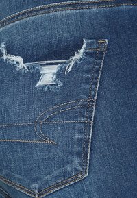 Jean en denim bleu foncé avec une poche arrière usée, présentant des bords effilochés et des coutures contrastantes. Le tissu a une apparence lisse et texturée.