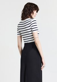 Donna con capelli castani corti e mossi, vestita con una maglietta bianca e blu a righe a maniche corte e pantaloni neri a vita alta, in posa di profilo.
