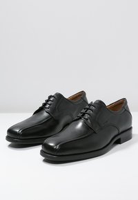 Zapatos de vestir de cuero negro con una textura suave, punta redondeada, diseño con cordones y un tacón bajo. Cuentan con costuras mínimas y un forro interior.