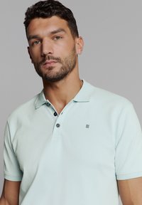 Licht mintgroene polo met zachte stof, voorzien van een drieknoopssluiting met contrasterende donkere knopen en een klein logo.