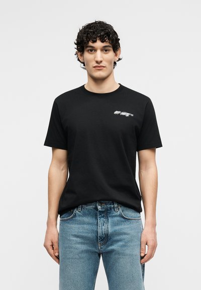 Νέος άνδρας με σγουρά μαλλιά φορώντας μαύρο T-shirt με το λογότυπο Off-White και μπλε τζιν, στέκεται μπροστά σε ένα απλό ανοιχτό γκρι φόντο.