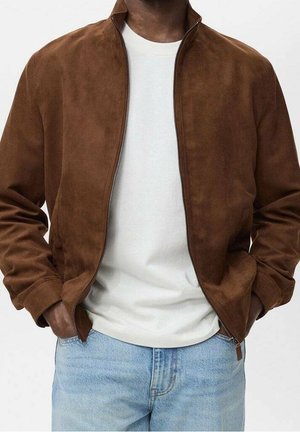 Homme portant une veste en daim marron dézippée par-dessus un t-shirt blanc et un jean bleu clair, les mains dans les poches de la veste.