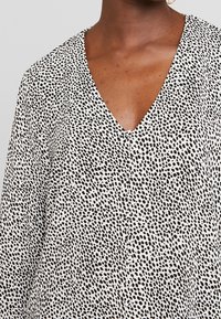 Schwarz-weiße gemusterte Bluse mit V-Ausschnitt und Knopfdetails, hergestellt aus einem leichten, strukturierten Stoff mit einem gesprenkelten Design.