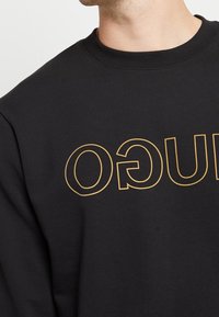 Czarna bluza z dużym złotym logo na przodzie. Materiał wydaje się gładki, z okrągłym dekoltem i ściągaczami na rękawach.