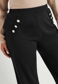 Svarta byxor i slät tyg har en bred elastisk midja och tre dekorativa silverknappar på sidan.